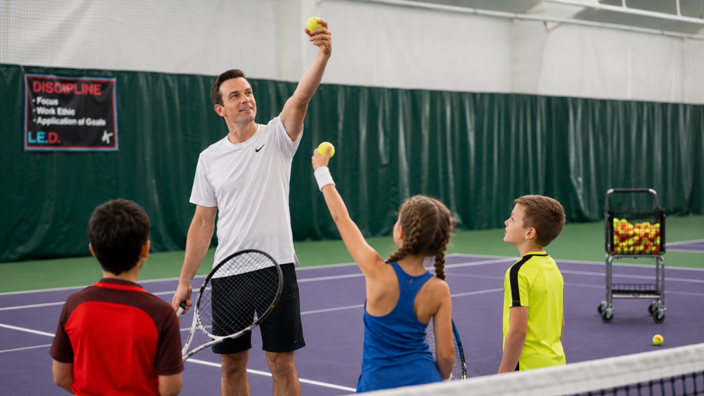 Tennis coach tossing a ball to two young players on an indoor court, racket in hand.