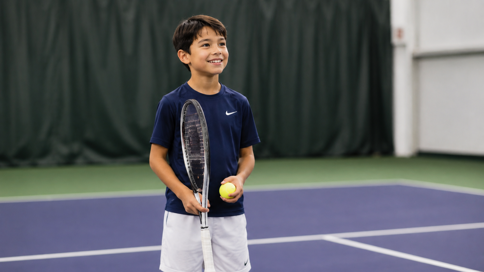 Young boy in a navy shirt holding a tennis racket and yellow ball on an indoor court, smiling at the camera.