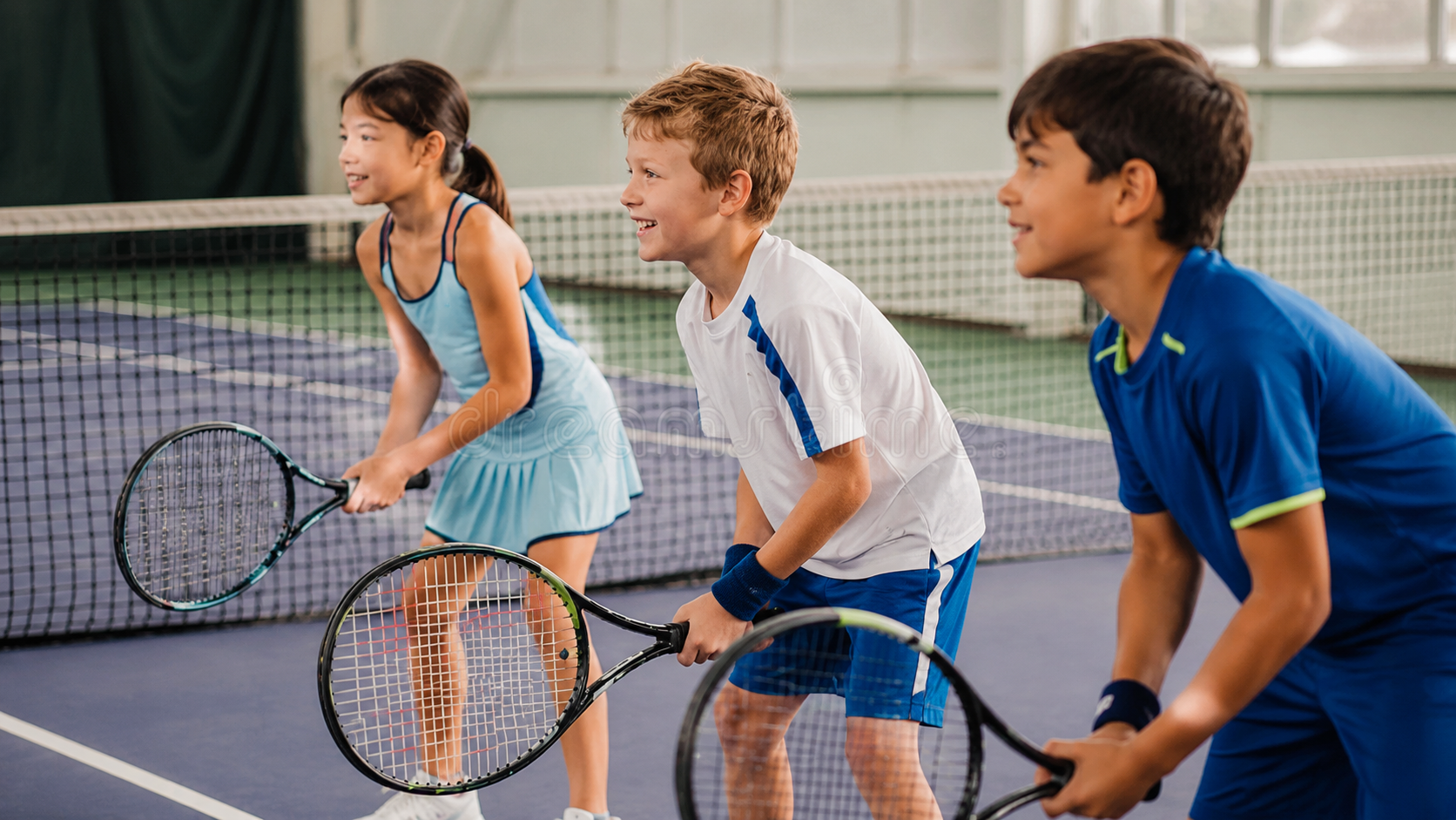 Three children in gym outfits stand with tennis rackets on an indoor court, ready to play.